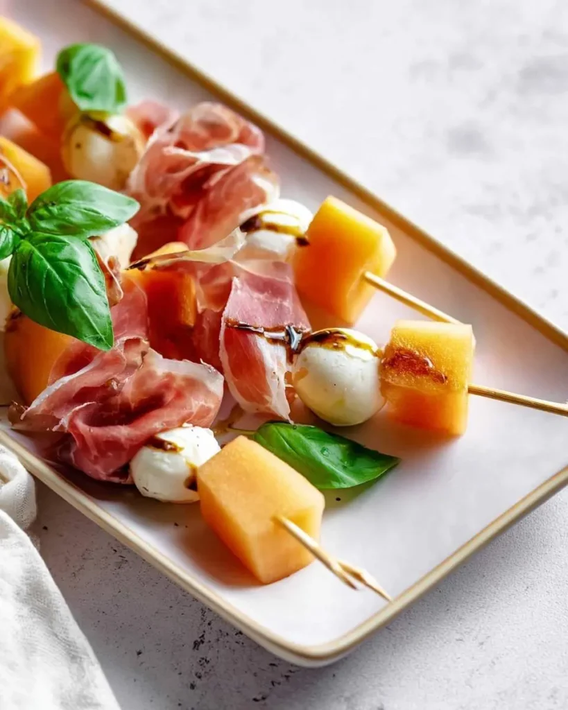 Close-up of fresh Melon Prosciutto Skewers with mozzarella, basil, and balsamic glaze on a white rectangular platter.