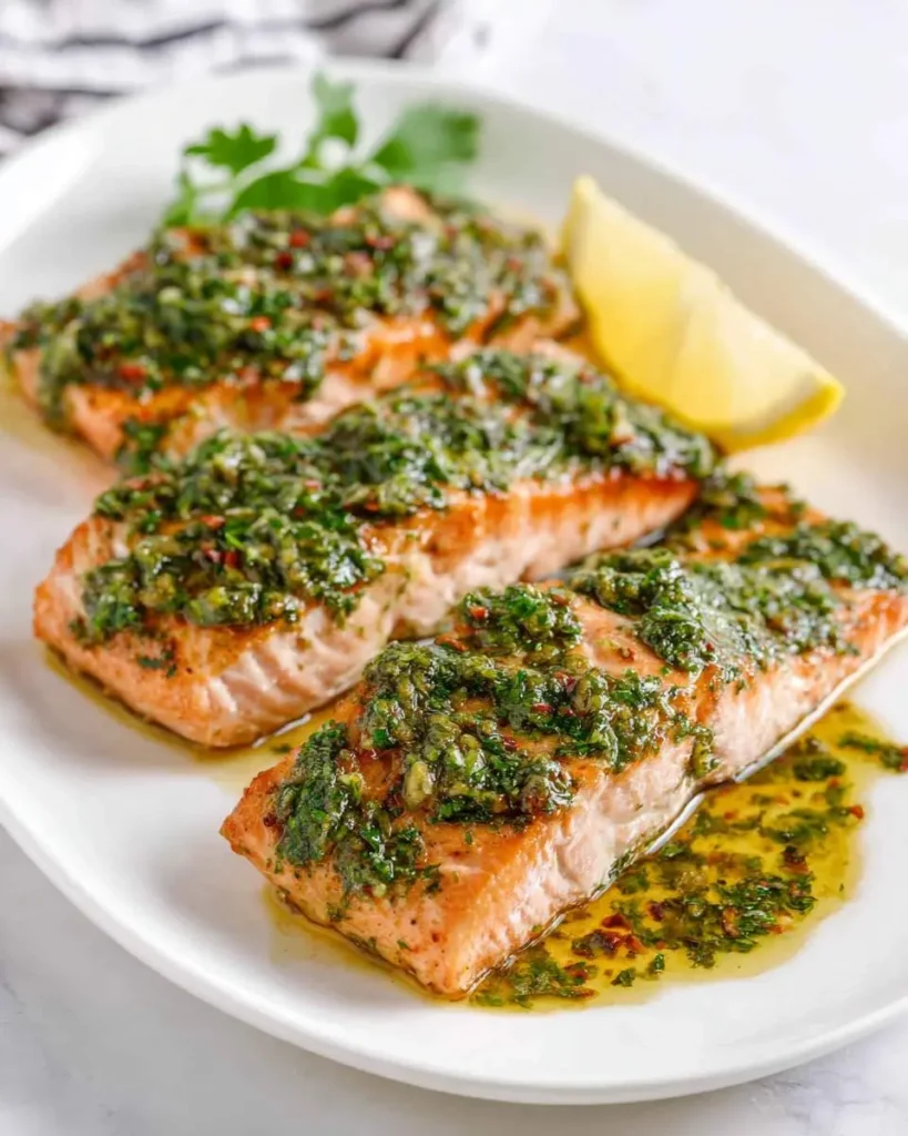 Three perfectly cooked salmon fillets topped with vibrant green chimichurri sauce and a lemon wedge on a white plate.