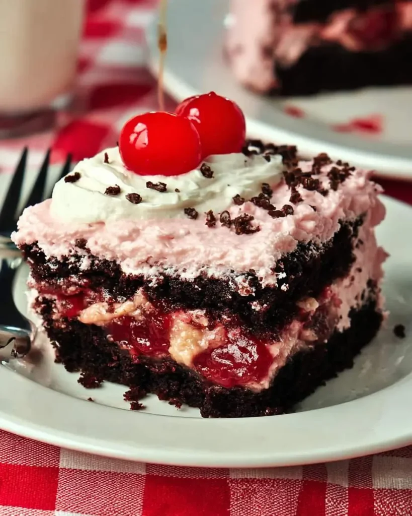 A slice of Cherry Dr Pepper Cake with chocolate layers, cherry filling, pink frosting, whipped cream, and maraschino cherries on top.