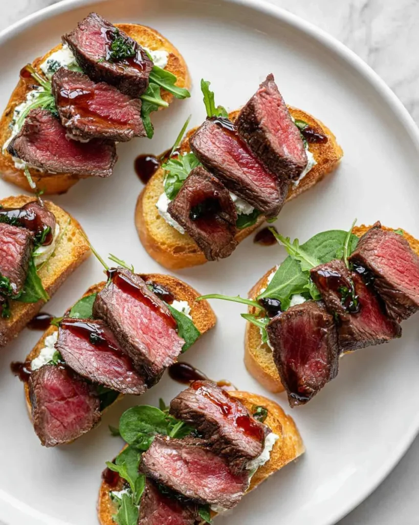 Six pieces of steak crostini arranged on a white plate, featuring toasted bread topped with creamy cheese, arugula, and sliced medium-rare beef drizzled with balsamic glaze.