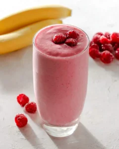 A tall glass of creamy banana raspberry smoothie topped with fresh raspberries, with bananas and berries in the background.